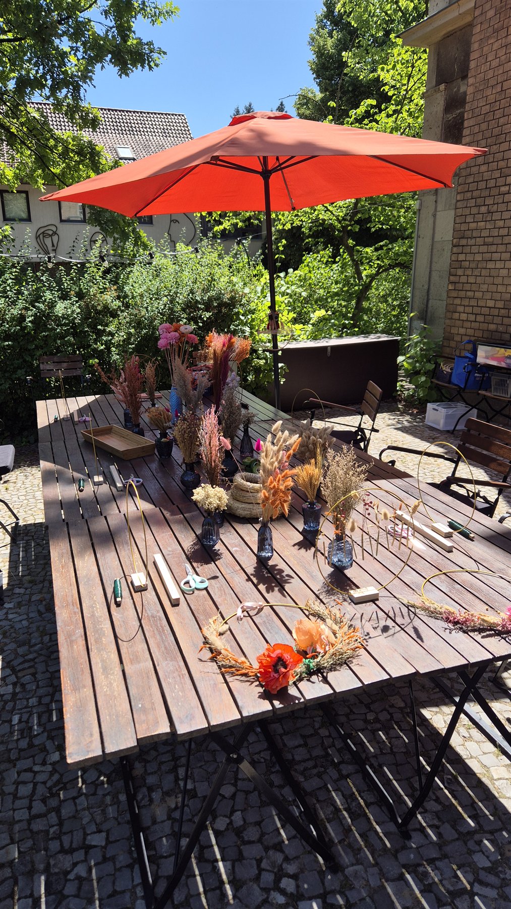 Vorbereiteter Workshoptisch mit Trockenblumen unter einem roten Sonnenschirm – ohne abgebildete Personen